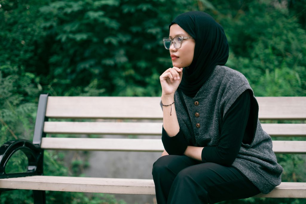 A woman wearing glasses and a black hijab sits on a park bench surrounded by greenery, resting her chin on her hand and looking into the distance.
