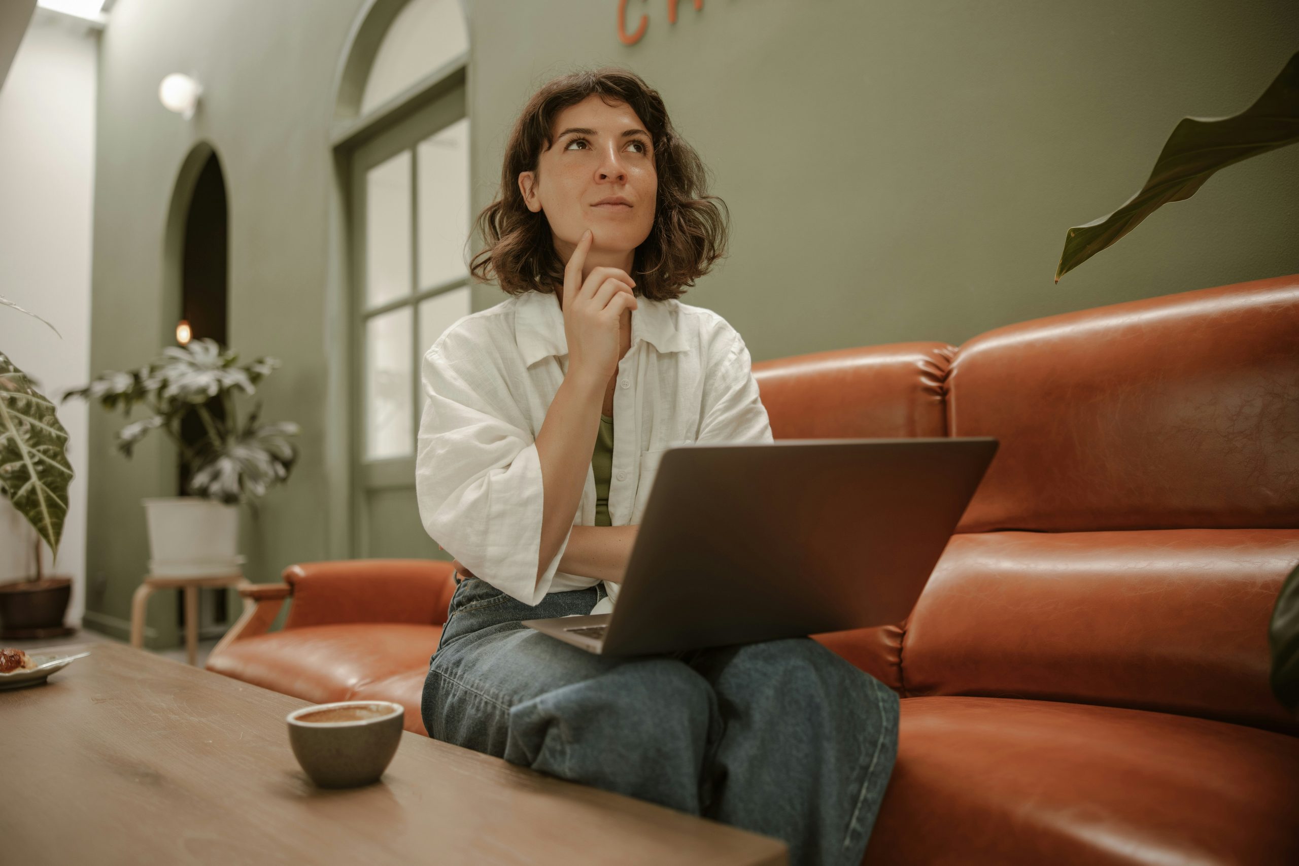 Woman sitting on a brown leather sofa with a laptop on her lap, looking upward with one finger resting against her chin.
