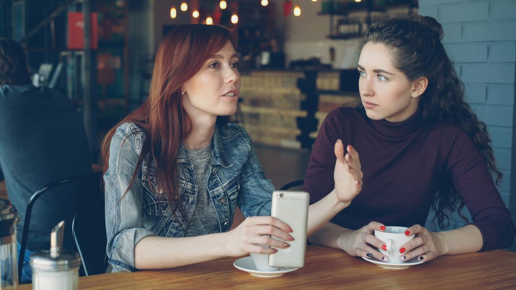 Two women talking at a café table, one gesturing while holding a phone and the other watching her with her hands around a cup.